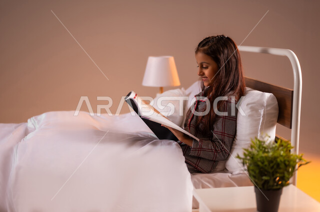 Relaxing and enjoying the calm atmosphere, reading interesting novels and stories, feeling comfortable and reassured, portrait of a smiling Saudi Arabian Gulf girl sitting in a comfortable bed holding a book in her hand, following a healthy lifestyle, colorful background