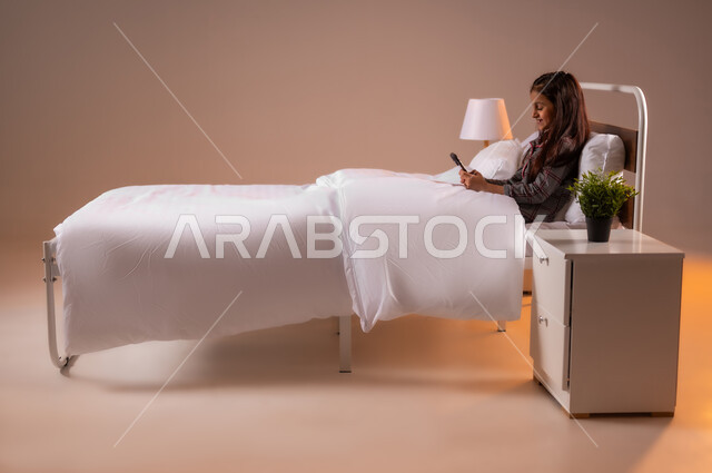 Gestures of integration and concentration, communicating with family and friends via social media, portrait of a Saudi Arabian Gulf girl sitting on a comfortable bed using a mobile phone, colorful background