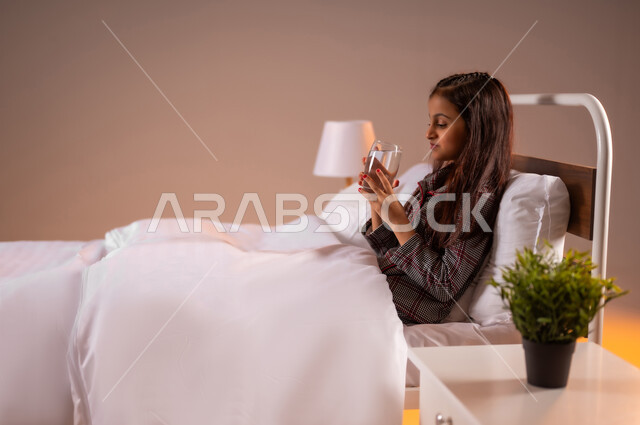 المداومة على شرب المياه، المحافظة على سلامة الجهاز المناعي للجسم، الشعور بالعطش والجفاف، بورتريه لفتاة عربية خليجية سعودية تجلس على سرير مريح تحمل بيدها كوب زجاجي تقوم بشرب الماء، خلفية ملونة