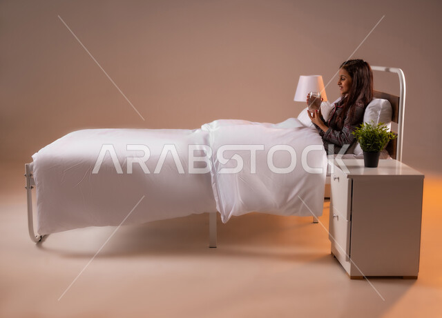 المحافظة على سلامة الجهاز المناعي للجسم، المداومة على شرب المياه، الشعور بالعطش والجفاف، بورتريه لفتاة عربية خليجية سعودية تجلس على سرير مريح تحمل بيدها كوب زجاجي تقوم بشرب الماء، خلفية ملونة