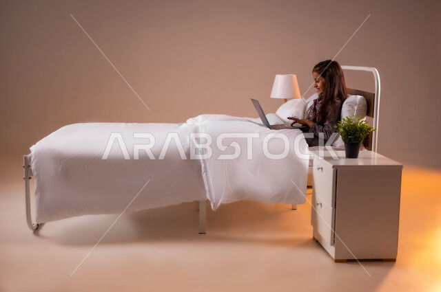 متابعة الدروس أونلاين، مفهوم التعلم عن بعد باستخدام الأجهزة التقنية الحديثة والتكنولوجيا، بورتريه لفتاة عربية خليجية سعودية مبتسمة تجلس على سرير مريح تحمل الحاسوب المحمول, ايماءات السرور والسعادة، خلفية ملونة