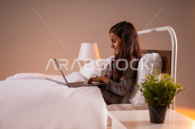 The concept of distance learning using modern technical devices and technology, portrait of a smiling Saudi Arabian Gulf girl sitting on a comfortable bed holding a laptop, following lessons online, gestures of pleasure and happiness, colorful background