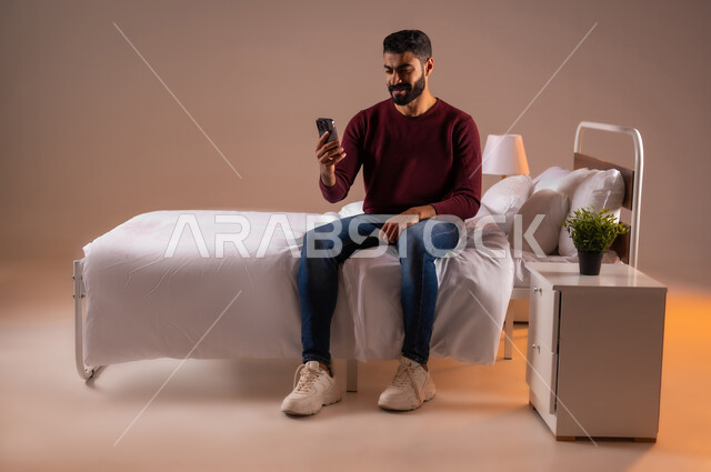 الدردشة والتواصل مع الاصدقاء بإيماءات السعادة والفرح، اجراء مكالمة فيديو، استخدام الأجهزة والتقنيات الحديثة، بورتريه لرجل عربي خليجي سعودي مبتسم يجلس على سرير مريح يمسك بيده الهاتف المحمول، تصفح الانترنت ومواقع التواصل الاجتماعي، صورة لكامل الجسم، خلفية ملونة