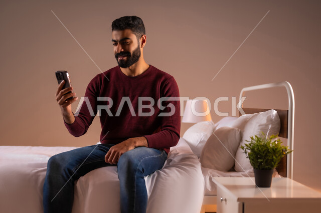 اجراء مكالمة فيديو، الدردشة والتواصل مع الاصدقاء بإيماءات السعادة والفرح، استخدام الأجهزة والتقنيات الحديثة، بورتريه لرجل عربي خليجي سعودي مبتسم يجلس على سرير مريح يمسك بيده الهاتف المحمول، تصفح الانترنت ومواقع التواصل الاجتماعي، خلفية ملونة