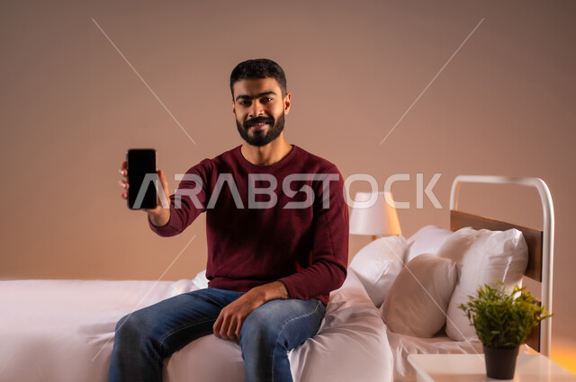 النظر الى الكاميرا بايماءات السعادة، عرض شاشة سوداء فارغة، تطور التكنولوجيا والأجهزة التقنية الحديثة، بورتريه لرجل عربي سعودي خليجي مبتسم يرتدي الزي الكاجوال يجلس على سرير مريح يحمل بيده الهاتف المحمول، خلفية ملونة