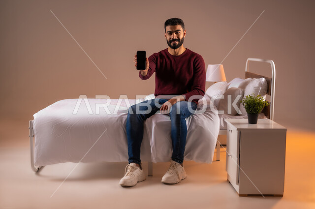 عرض شاشة سوداء فارغة، النظر الى الكاميرا بايماءات السعادة، تطور التكنولوجيا والأجهزة التقنية الحديثة، بورتريه لرجل عربي سعودي خليجي مبتسم يرتدي الزي الكاجوال يجلس على سرير مريح يحمل بيده الهاتف المحمول، صورة لكامل الجسم، خلفية ملونة