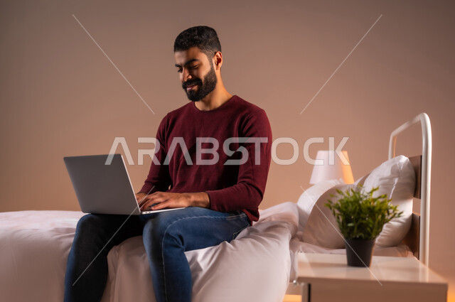 عقد إجتماع عمل عبر الإنترنت، تصفح برامج التواصل الاجتماعي, إستخدام الحاسوب المحمول لإنجاز المهام، بورتريه لرجل عربي خليجي سعودي يرتدي الزي الكاجوال يحمل جهاز اللابتوب يجلس على سريره في غرفة النوم، تطور التكنولوجيا والأجهزة التقنية، خلفية ملونة