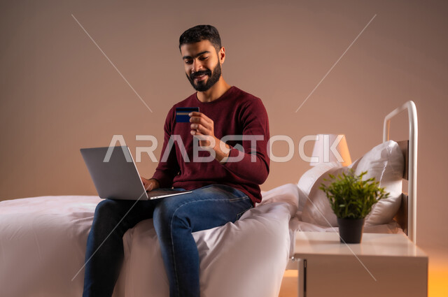 استخدام البطاقة البنكية الالكترونية، مفهوم الدفع والتسديد عن بعد عبر تطبيقات اللابتوب، تطور التكنولوجيا والأجهزة التقنية، بورتريه لرجل عربي سعودي خليجي يرتدي الزي الكاجوال يجلس على سرير مريح يعمل على جهاز الحاسوب المحمول، خلفية ملونة