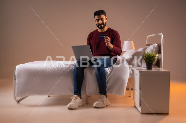 مفهوم الدفع والتسديد عن بعد عبر تطبيقات اللابتوب، استخدام البطاقة البنكية الالكترونية، تطور التكنولوجيا والأجهزة التقنية، بورتريه لرجل عربي سعودي خليجي يرتدي الزي الكاجوال يجلس على سرير مريح يعمل على جهاز الحاسوب المحمول، صورة لكامل الجسم، خلفية ملونة