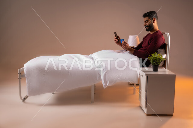 التسوق الالكتروني عبر الانترنت، مفهوم الدفع والتسديد عن بعد عبر تطبيقات الجوال، استخدام البطاقة البنكية الالكترونية، بورتريه لرجل عربي سعودي خليجي يرتدي ازي الكاجوال مستلقي على سرير مريح يحمل الهاتف المحمول، خلفية ملونة