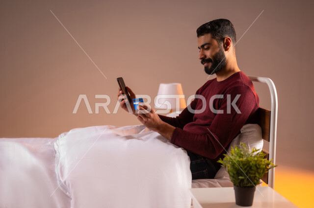 التسوق الالكتروني عبر الانترنت، مفهوم الدفع والتسديد عن بعد عبر تطبيقات الجوال، استخدام البطاقة البنكية الالكترونية، بورتريه لرجل عربي سعودي خليجي يرتدي ازي الكاجوال مستلقي على سرير مريح يحمل الهاتف المحمول، خلفية ملونة