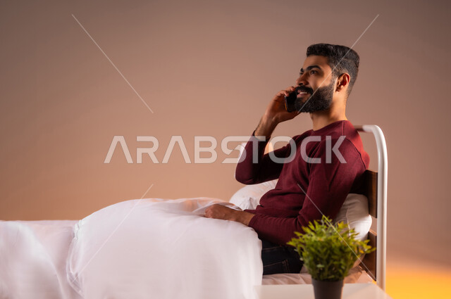 إيماءات الفرح والسرور، إجراء مكالمة هاتفية مع الأهل والأصدقاء، بورتريه لرجل عربي سعودي خليجي مبتسم يرتدي الزي الكاجوال يستلقي على سرير مريح يحمل بيده الهاتف المحمول، تطور التكنولوجيا والأجهزة التقنية، خلفية ملونة