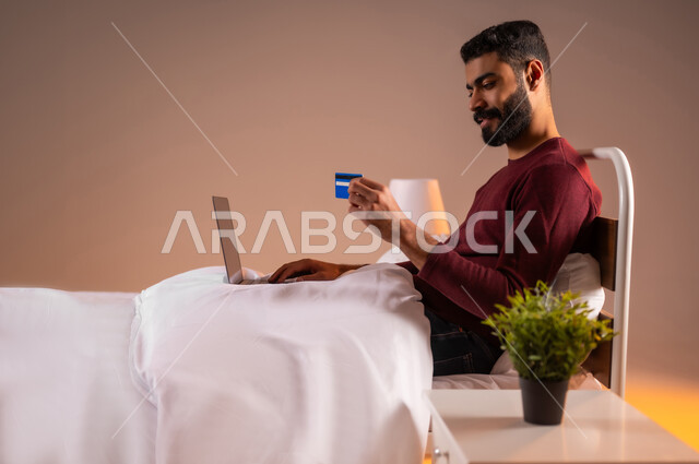 مفهوم الدفع والتسديد عن بعد عبر تطبيقات اللابتوب، استخدام البطاقة البنكية الالكترونية، تطور التكنولوجيا والأجهزة التقنية، بورتريه لرجل عربي سعودي خليجي يرتدي الزي الكاجوال مستلقي على سرير مريح يعمل على جهاز الحاسوب المحمول، خلفية ملونة