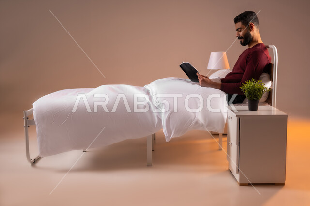 هواية المطالعة والاستكشاف، قضاء وقت الفراغ بالتعلم والثقافة، مفهوم التثقيف والبحث العلمي، بورتريه لشاب عربي خليجي سعودي مبتسم يرتدي الزي الكاجوال مستلقي على سرير مريح يقوم قراءة كتاب, خلفية ملونة