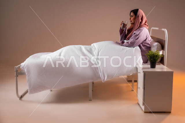 Feeling thirsty and dehydrated, maintaining the health of the body's immune system, drinking water regularly, portrait of a veiled Saudi Arabian Gulf woman sitting on a comfortable bed holding a glass cup in her hand drinking water, colorful background