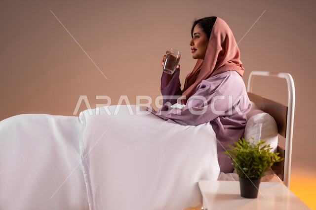 المحافظة على سلامة الجهاز المناعي للجسم، المداومة على شرب المياه، الشعور بالعطش والجفاف، بورتريه لامرأة عربية خليجية سعودية محجبة تجلس على سرير مريح تحمل بيدها كوب زجاجي تقوم بشرب الماء، خلفية ملونة