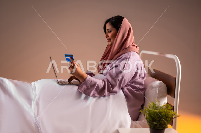 استخدام الاجهزة التقنية الحديثة، مفهوم الدفع عن بعد عبر تطبيقات اللابتوب، استخدام البطاقة البنكية الالكترونية، بورتريه لإمرأة عربية سعودية خليجية محجبة مبتسمة تجلس على سرير تقوم بالتسوق عبر جهاز الحاسوب المحمول، خلفية ملونة