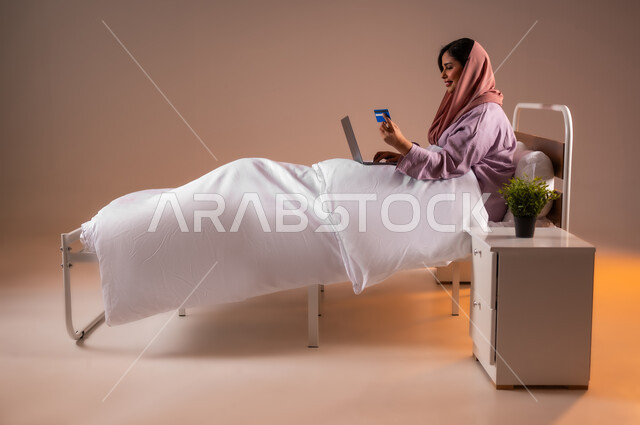 مفهوم الدفع عن بعد عبر تطبيقات اللابتوب، استخدام البطاقة البنكية الالكترونية، بورتريه لإمرأة عربية سعودية خليجية محجبة مبتسمة تجلس على سرير تقوم بالتسوق عبر جهاز الحاسوب المحمول، استخدام الاجهزة التقنية الحديثة، خلفية ملونة