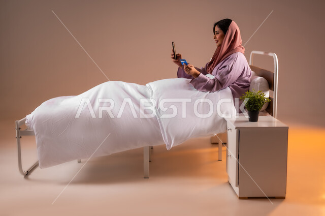 مفهوم الدفع عن بعد عبر تطبيقات الجوال، استخدام البطاقة البنكية الالكترونية، التكنولوجيا الحديثة والتقنيات المتطورة، إمرأة عربية سعودية خليجية محجبة مبتسمة تجلس على سرير مريح تقوم بالتسوق باستعمال الهاتف المحمول، خلفية ملونة