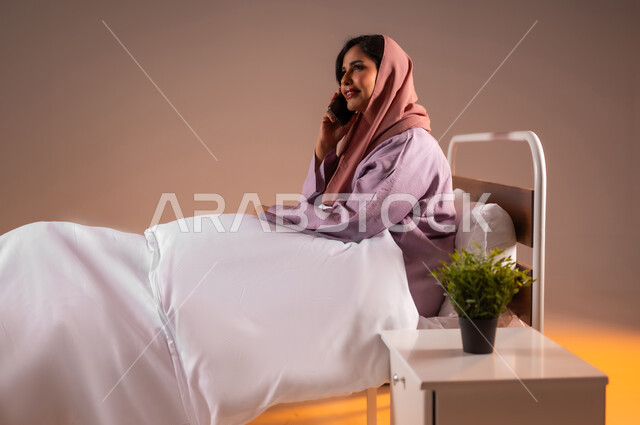 محادثات وحوارات ودية، ايماءات السعادة و السرور، التواصل مع الأهل والأصدقاء باستخدام الاجهزة التقنية الحديثة، بورتريه لامراة عربية خليجية سعودية محجبة مبتسمة تجلس على سرير مريح تحمل بيدها الهاتف المحمول تقوم باجراء مكالمة هاتفية، خلفية ملونة