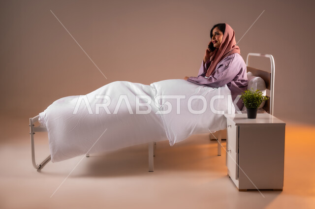 ايماءات السعادة و السرور، محادثات وحوارات ودية، التواصل مع الأهل والأصدقاء باستخدام الاجهزة التقنية الحديثة، بورتريه لامراة عربية خليجية سعودية محجبة مبتسمة تجلس على سرير مريح تحمل بيدها الهاتف المحمول تقوم باجراء مكالمة هاتفية، خلفية ملونة