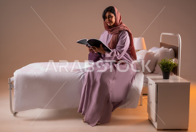 الشعور بالراحة والطمأنينة, قراءة الروايات والقصص الممتعة، بورتريه لسيدة عربية خليجية سعودية مبتسمة تجلس في سرير مريح تمسك بيدها الكتاب، الاسترخاء والاستمتاع بالأجواء الهادئة، اتباع أسلوب حياة صحي، صورة لكامل الجسم، خلفية ملونة