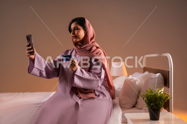 استخدام البطاقة البنكية الالكترونية، التكنولوجيا الحديثة والتقنيات المتطورة، إمرأة عربية سعودية خليجية محجبة مبتسمة تجلس على سرير مريح تقوم بالتسوق باستعمال الهاتف المحمول، مفهوم الدفع عن بعد عبر تطبيقات الجوال، خلفية ملونة