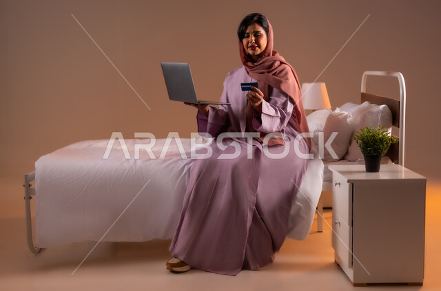 استخدام البطاقة البنكية الالكترونية، مفهوم الدفع عن بعد عبر تطبيقات اللابتوب، بورتريه لإمرأة عربية سعودية خليجية محجبة مبتسمة تجلس على سرير تقوم بالتسوق عبر جهاز الحاسوب المحمول، استخدام الاجهزة التقنية الحديثة، صورة لكامل الجسم، خلفية ملونة