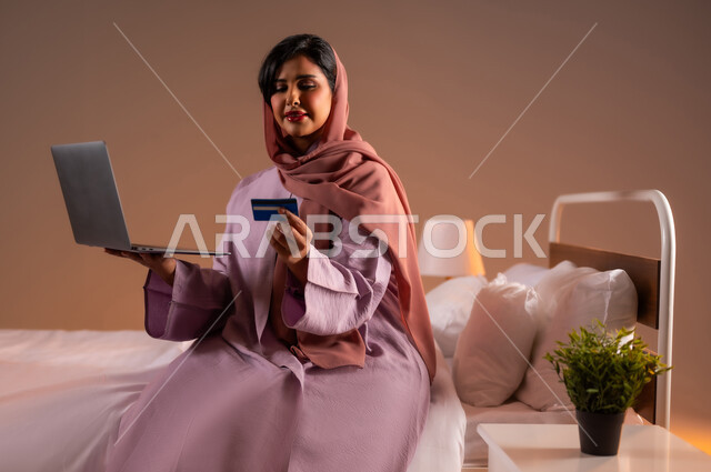 استخدام البطاقة البنكية الالكترونية، مفهوم الدفع عن بعد عبر تطبيقات اللابتوب، بورتريه لإمرأة عربية سعودية خليجية محجبة مبتسمة تجلس على سرير مريح تقوم بالتسوق عبر جهاز الحاسوب المحمول، إيماءات السعادة والسرور، استخدام الاجهزة التقنية الحديثة، خلفية ملونة