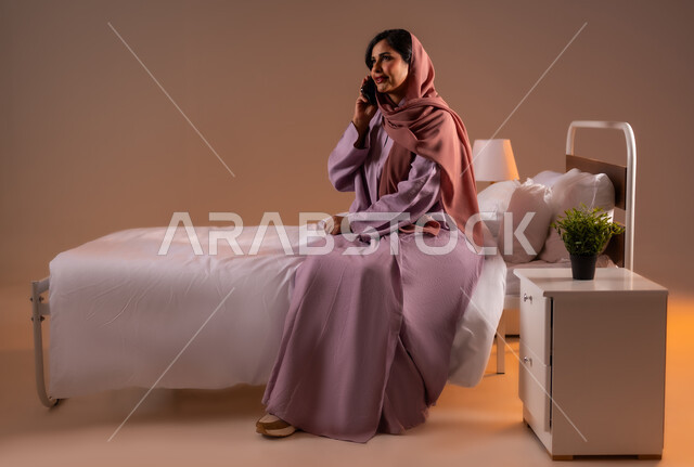 التواصل مع الأهل والأصدقاء باستخدام الاجهزة التقنية الحديثة، محادثات وحوارات ودية، ايماءات السعادة و السرور، بورتريه لامراة عربية خليجية سعودية محجبة مبتسمة تجلس على سرير مريح تحمل بيدها الهاتف المحمول تقوم باجراء مكالمة هاتفية، صورة لكامل الجسم، خلفية ملونة