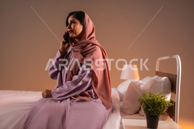 Friendly conversations and dialogues, gestures of happiness and pleasure, communicating with family and friends using modern technical devices, portrait of a smiling veiled Saudi Arabian Gulf woman sitting on a comfortable bed holding a mobile phone in her hand making a phone call, colorful background
