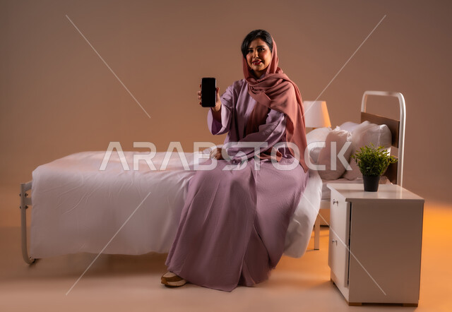 عرض شاشة سوداء فارغة، النظر الى الكاميرا بايماءات السعادة، تطور التكنولوجيا والأجهزة التقنية الحديثة، بورتريه لامرأة عربية سعودية خليجية مبتسمة ترتدي الثوب العباءة تجلس على سرير مريح تحمل بيدها الهاتف المحمول، صورة لكامل الجسم، خلفية ملونة