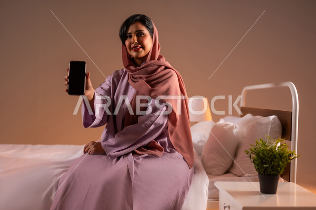 عرض شاشة سوداء فارغة، النظر الى الكاميرا بايماءات السعادة، تطور التكنولوجيا والأجهزة التقنية الحديثة، بورتريه لامرأة عربية سعودية خليجية مبتسمة ترتدي الثوب العباءة تجلس على سرير مريح تحمل بيدها الهاتف المحمول، خلفية ملونة