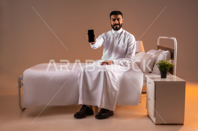 Looking at the camera with gestures of happiness, development of technology and modern technical devices, portrait of a smiling Saudi Arabian Gulf man wearing traditional dress sitting on a comfortable bed displaying a blank black screen on a mobile phone, full body image, sand background