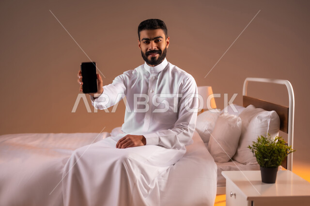 تطور التكنولوجيا والأجهزة التقنية الحديثة، بورتريه لرجل عربي سعودي خليجي مبتسم يرتدي الثوب التقليدي يجلس على سرير مريح يقوم بعرض شاشة سوداء فارغة على الهاتف المحمول، خلفية ملونة