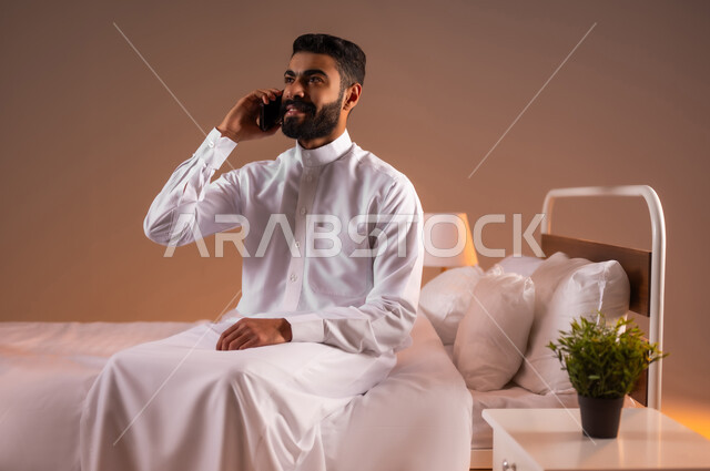 Gestures of joy and happiness, making a phone call with family and friends, portrait of a smiling Saudi Arabian Gulf man wearing traditional thobe sitting on a comfortable bed holding a mobile phone in his hand, development of technology and technical devices, colorful background