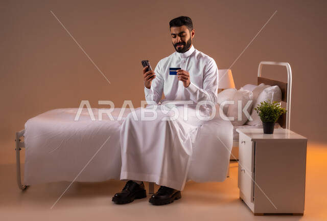 التسوق الالكتروني عبر الانترنت، مفهوم الدفع والتسديد عن بعد عبر تطبيقات الجوال، استخدام البطاقة البنكية الالكترونية، بورتريه لرجل عربي سعودي خليجي يرتدي الثوب التقليدي يجلس على سرير مريح يحمل الهاتف المحمول، صورة لكامل الجسم، خلفية ملونة