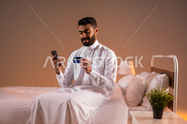 التسوق الالكتروني عبر الانترنت، مفهوم الدفع والتسديد عن بعد عبر تطبيقات الجوال، استخدام البطاقة البنكية الالكترونية، بورتريه لرجل عربي سعودي خليجي يرتدي الثوب التقليدي يجلس على سرير مريح يحمل الهاتف المحمول، خلفية ملونة
