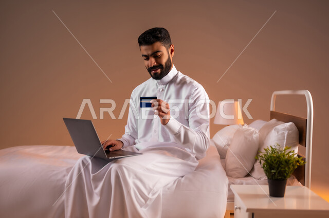استخدام البطاقة البنكية الالكترونية، مفهوم الدفع والتسديد عن بعد عبر تطبيقات اللابتوب، تطور التكنولوجيا والأجهزة التقنية، بورتريه لرجل عربي سعودي خليجي يرتدي الثوب التقليدي يجلس على سرير مريح يعمل على جهاز الحاسوب المحمول، خلفية ملونة