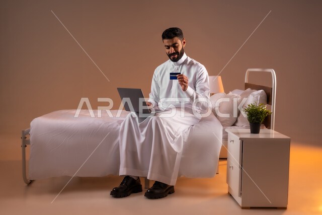 مفهوم الدفع والتسديد عن بعد عبر تطبيقات اللابتوب، استخدام البطاقة البنكية الالكترونية، تطور التكنولوجيا والأجهزة التقنية، بورتريه لرجل عربي سعودي خليجي يرتدي الثوب التقليدي يجلس على سرير مريح يعمل على جهاز الحاسوب المحمول، صورة لكامل الجسم، خلفية ملونة