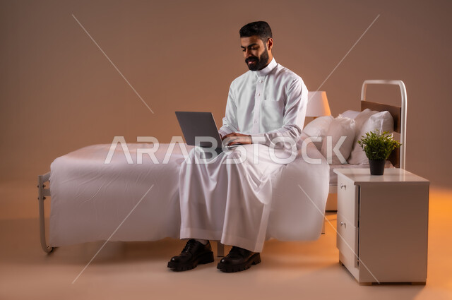 عقد إجتماع عمل عبر الإنترنت، تصفح برامج التواصل الاجتماعي, إستخدام الحاسوب المحمول لإنجاز المهام، بورتريه لرجل عربي خليجي سعودي يرتدي الثوب التقليدي يحمل جهاز اللابتوب يجلس على سريره في غرفة النوم، تطور التكنولوجيا والأجهزة التقنية