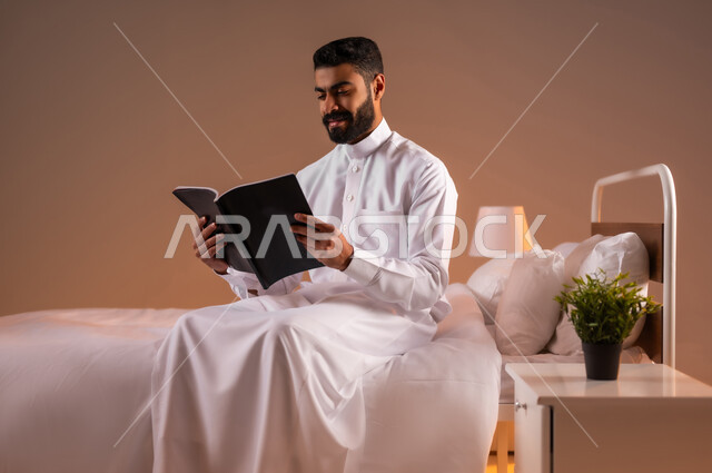 قضاء وقت الفراغ بالتعلم والثقافة، مفهوم التثقيف والبحث العلمي، بورتريه لشاب عربي خليجي سعودي مبتسم يرتدي الثوب التقليدي يجلس على سرير مريح يقوم قراءة كتاب, هواية المطالعة والاستكشاف، خلفية ملونة