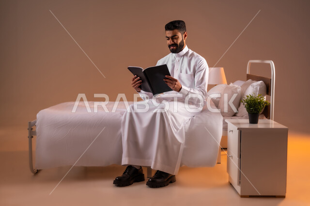 مفهوم التثقيف والبحث العلمي، قضاء وقت الفراغ بالتعلم والثقافة، بورتريه لشاب عربي خليجي سعودي مبتسم يرتدي الثوب التقليدي يجلس على سرير مريح يقوم قراءة كتاب, هواية المطالعة والاستكشاف، صورة لكامل الجسم، خلفية ملونة