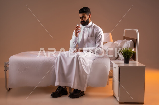 المحافظة على سلامة الجهاز المناعي للجسم، المداومة على شرب المياه، الشعور بالعطش والجفاف، بورتريه لرجل عربي خليجي سعودي يرتدي الثوب التقليدي يجلس على سرير مريح يحمل بيده كوب زجاجي يقوم بشرب الماء، صورة لكامل الجسم، خلفية ملونة