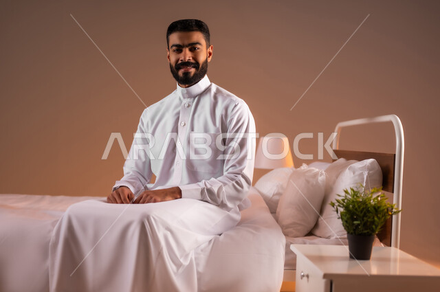 The concept of elegance and masculinity, looking at the camera with gestures of self-confidence, expressions of pleasure and happiness, interest in outward appearance, portrait of a Saudi Arabian Gulf man wearing traditional thobe sitting on a comfortable bed, colorful background