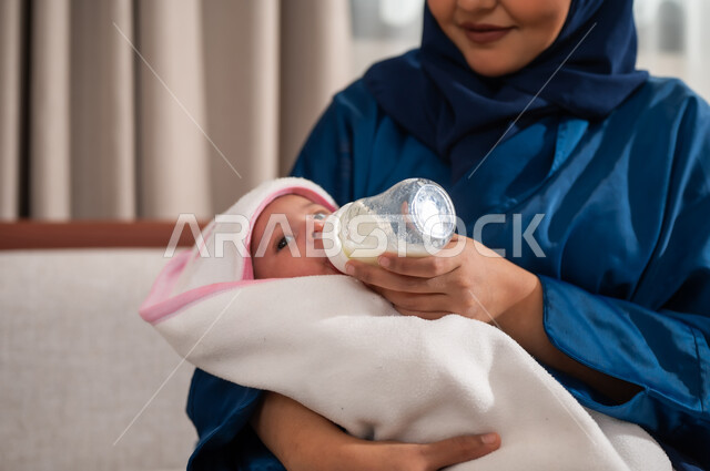 دور و حنان الأم في رعايه أبنائها, قضاء أوقات ممتعة مع الأطفال، الأسرة وتحمل المسؤولية،  صورة مقربة لسيدة عربيه خليجيه سعوديه ترتدي العباءة تحمل ابنتها وتقوم بإعطاءها الحليب الصناعي من عبوه الرضاعه, مفهوم الأمومه والرضاعه الصناعيه, ايماءات تدل على الحب والحنان