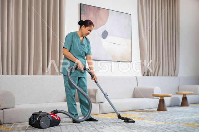 المهام والأعمال المنزلية الروتينية، عاملة نظافة ترتدي اليونيفورم الخاص بالعمل تقوم بتنظيف البيت بالمكنسة الكهربائية، استقدام العمالة الاجنبية المنزليه المدربة، مهن ووظائف نسائية، جودة الخدمات في السعودية