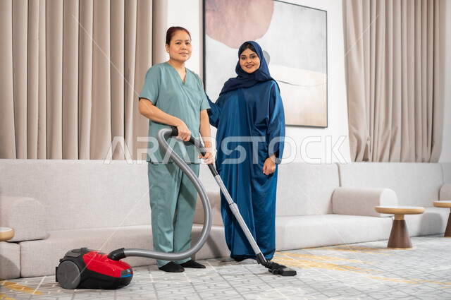 استقدام العمالة الخارجية المرخصة والمؤهلة، توفير خدمات العناية بالمنزل والنظافة والترتيب، النظر الى الكاميرا بايماءات السعادة والرضا عن جودة خدمة العاملة، سيدة عربية خليجية سعودية تعطي الارشادات والتوجيهات لخادمة فلبينية تقوم باستخدام المكنسة الكهربائية