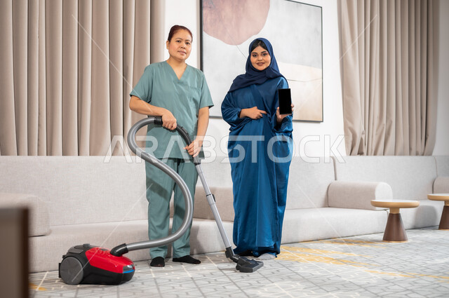 الخدمات الإلكترونيه المتقدمه لدعم وتوفير الخادمات المرخصة والمؤهله والمدربة، النظر الى الكاميرا بايماءات الرضا عن جودة خدمة العاملة، سيدة عربية خليجية سعودية تحمل بيدها هاتف مححمول بشاشة سوداء فارغة، خادمة فلبينية تقوم باستخدام المكنسة الكهربائية