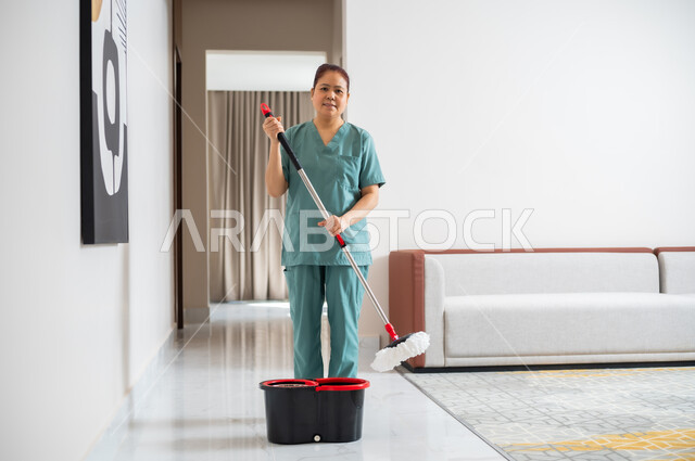 عاملة نظافة تنظر الى الكاميرا ترتدي اليونيفورم الخاص بالعمل تقوم بمسح وتنظيف البيت، استقدام العمالة الاجنبية المنزليه المدربة، الاهتمام بالتنظيف والقيام بالمهام الروتينة، مهن ووظائف نسائية، جودة الخدمات في السعودية